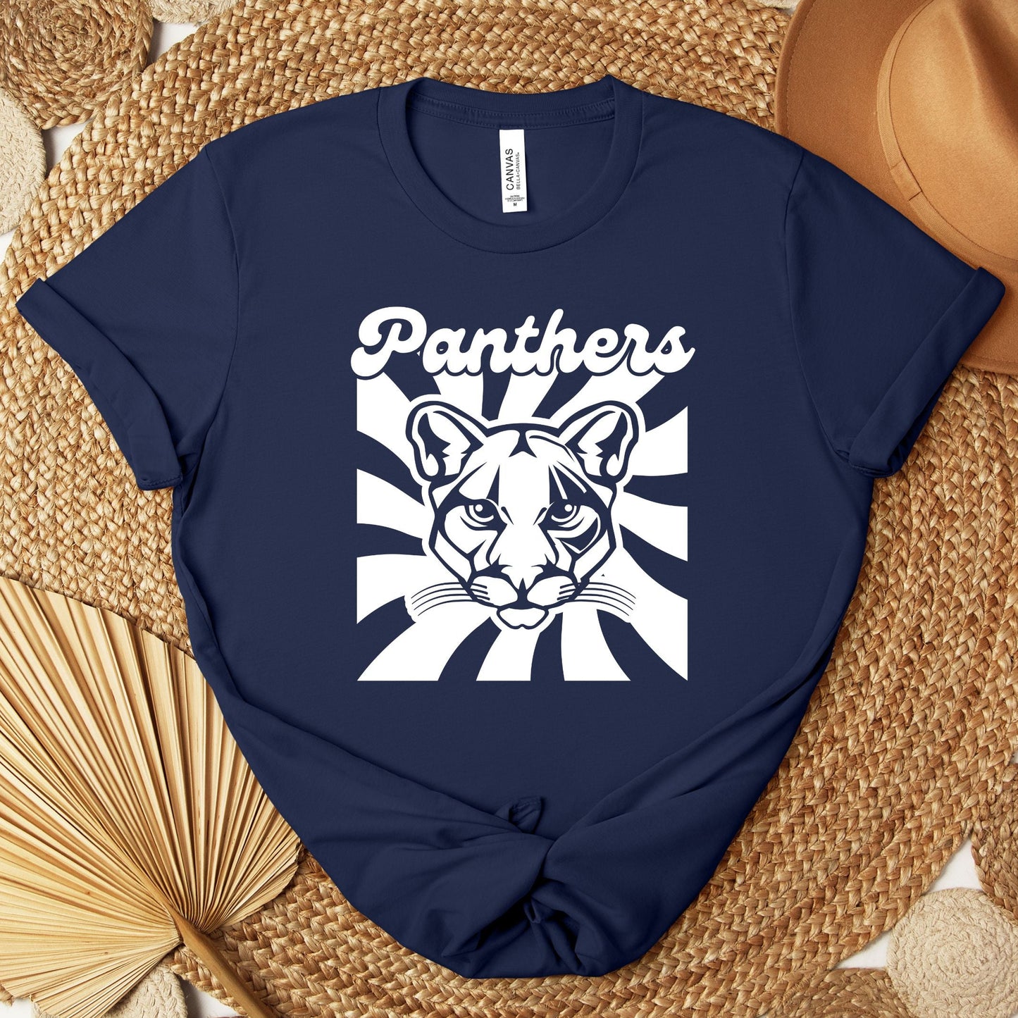 A navy blue short-sleeve T-shirt laid flat on a woven mat, featuring a white graphic of a panther face with the word 'Panthers' above it. A tan hat and dried palm leaves are placed around the shirt as styling props.