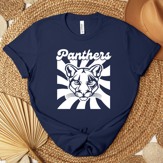 A navy blue short-sleeve T-shirt laid flat on a woven mat, featuring a white graphic of a panther face with the word 'Panthers' above it. A tan hat and dried palm leaves are placed around the shirt as styling props.